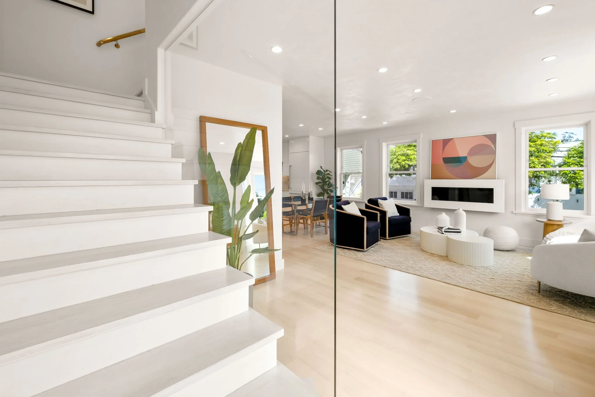 A bright modern living room with white walls, large windows, and light wooden flooring. The room features a white sofa, black armchairs, a fireplace, a large abstract painting, and decorative objects. To the left, a mirror leans against the wall next