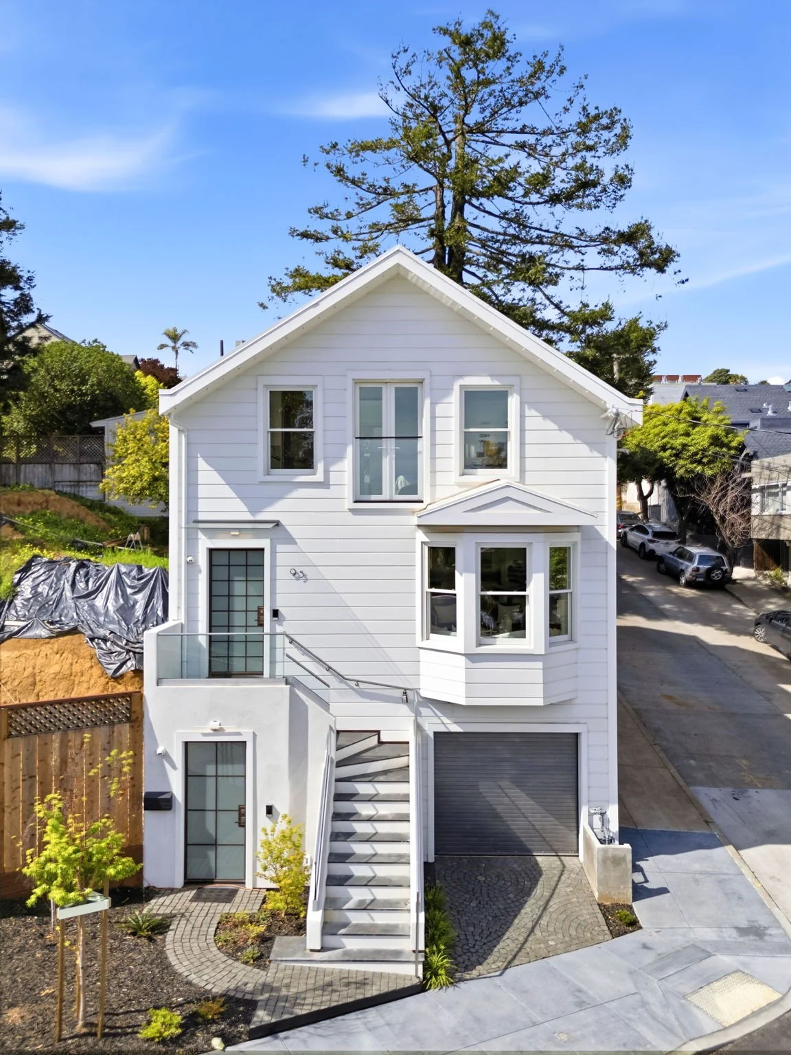 A elevated white house with a garage, multiple windows, and a staircase leading to the front door, surrounded by small landscaped areas and parked cars nearby.