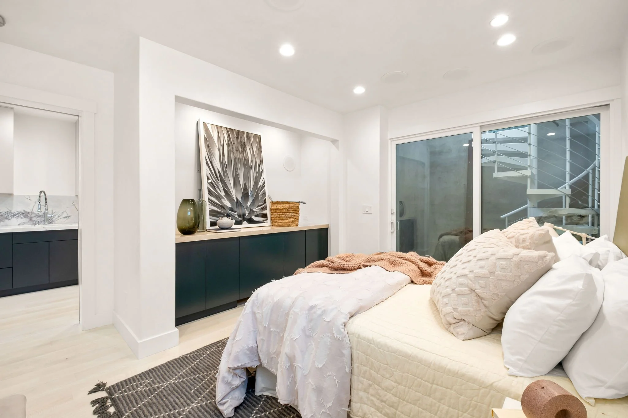 Modern bedroom with white walls, a bed with cream and white bedding, large pillows, a sliding glass door leading outside, and a sideboard decorated with artwork, vases, and a basket.
