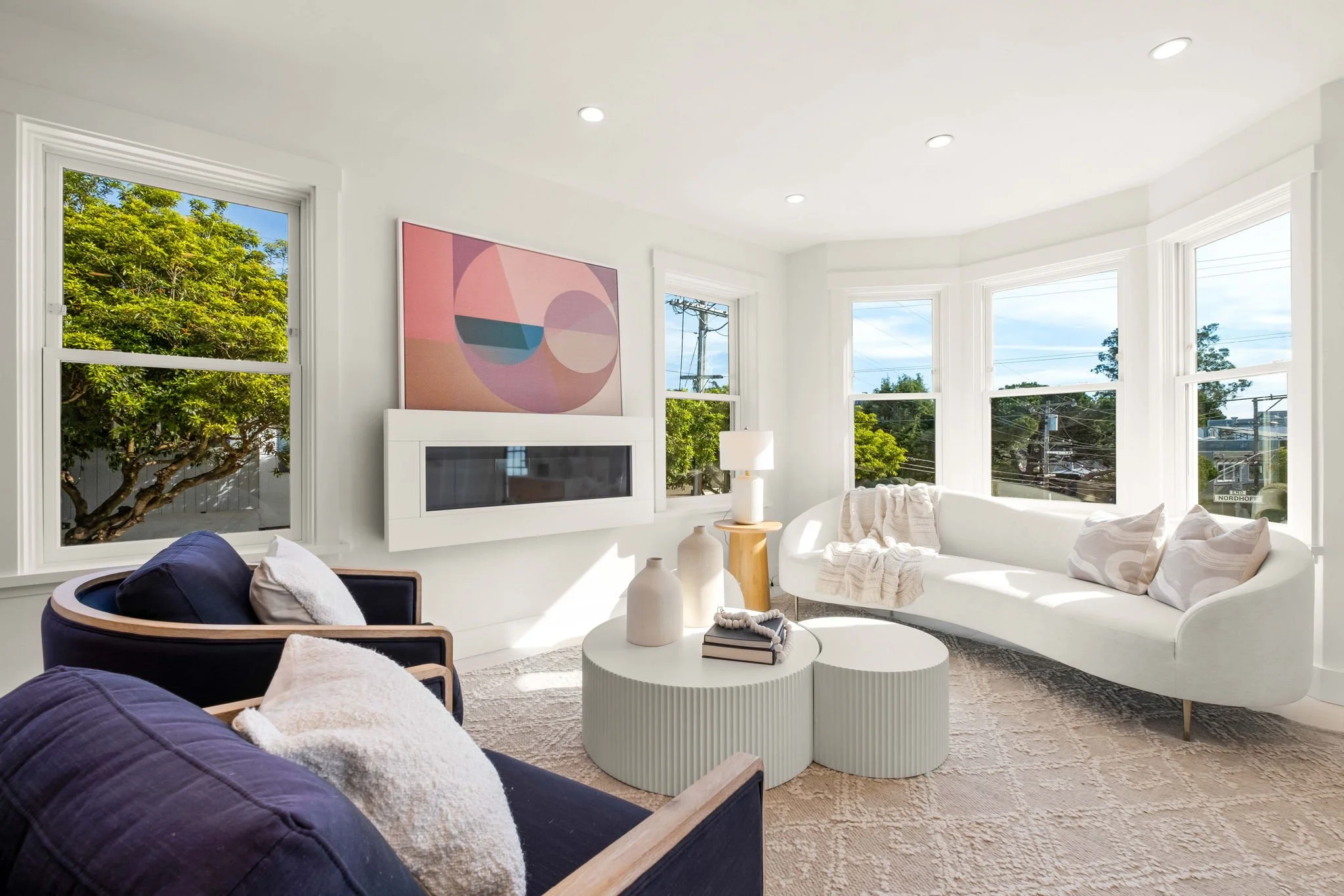 Bright living room with white walls, large windows, modern white curved sofa with pillows, dark armchair with pillows, round white coffee tables with vases and books, abstract pink art hanging above a white electric fireplace, light-colored rug, tabl