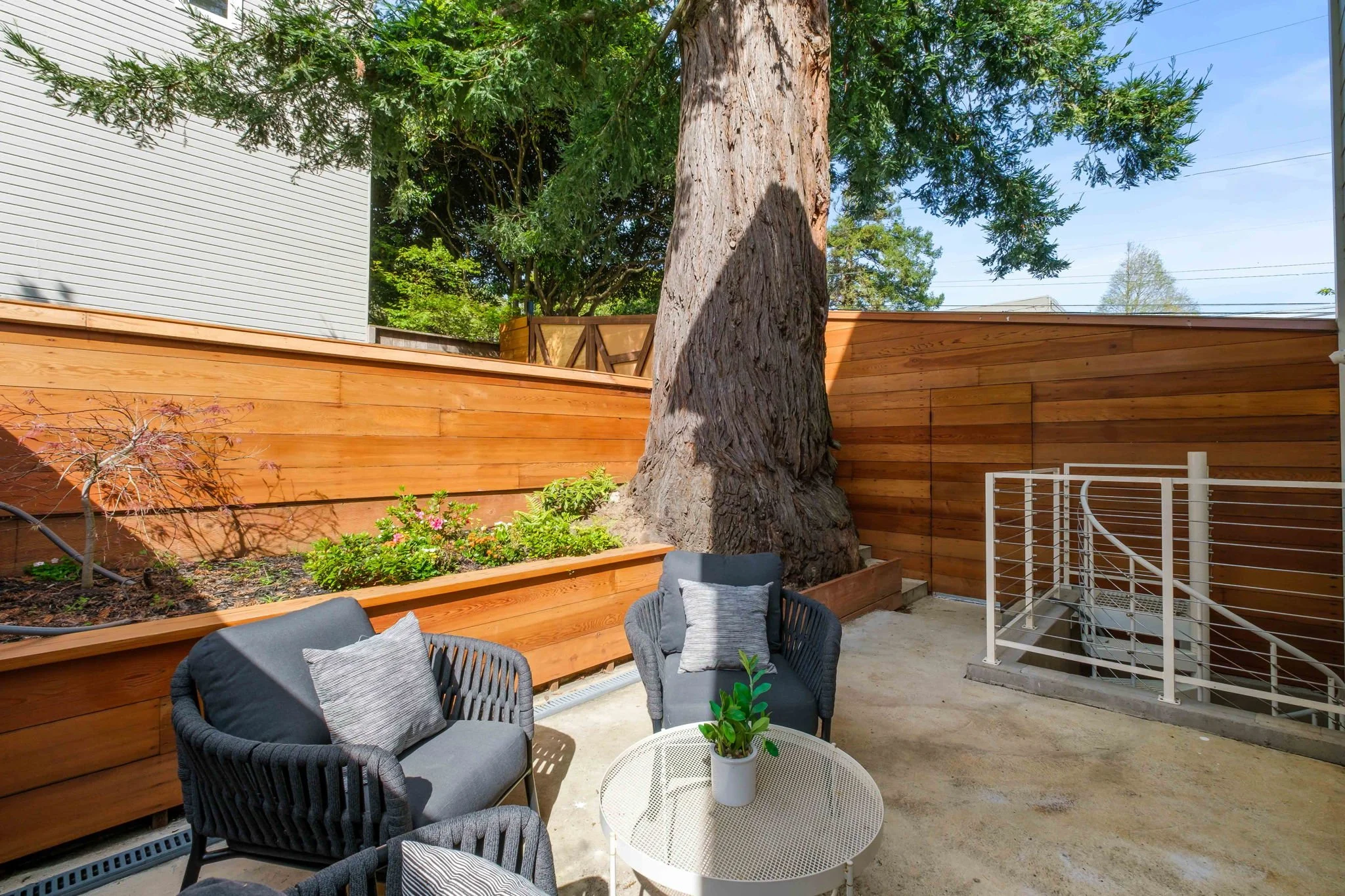 A backyard patio with a large tree and a raised wooden garden bed with plants and flowers. There are two gray outdoor chairs with cushions and a white round table with a potted plant. Wooden privacy fencing surrounds the area, and a staircase with a 