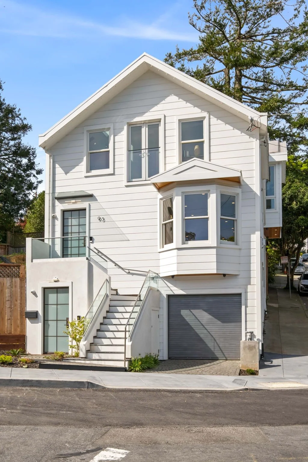 A modern white multi-story house with stairs leading to a second-floor entrance, bay windows, and a garage on the ground level, with trees and a street in the background.