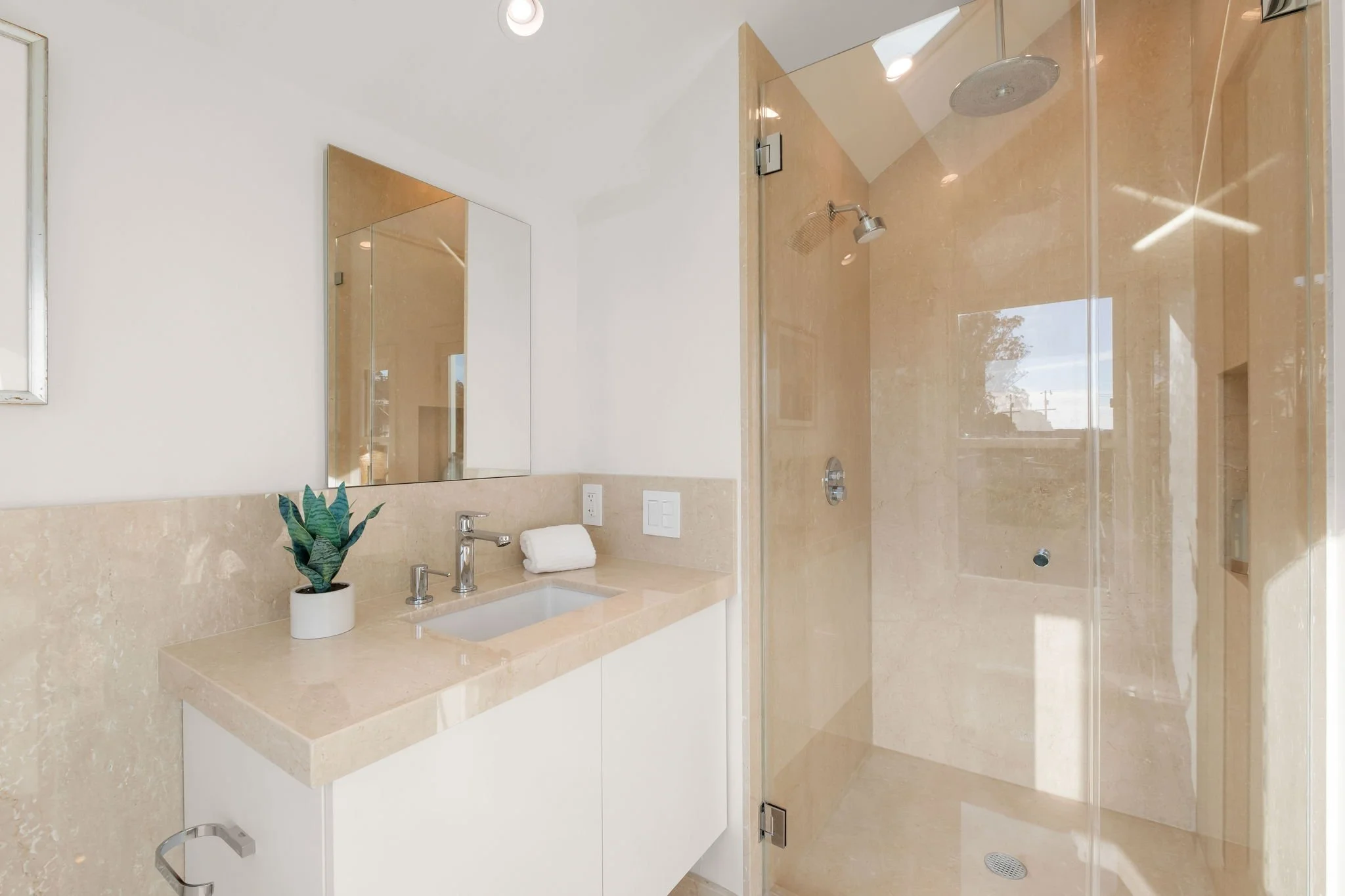 Modern bathroom with a beige marble vanity top, a rectangular mirror, a potted plant, and a glass-enclosed walk-in shower with beige marble walls and a rainfall showerhead.