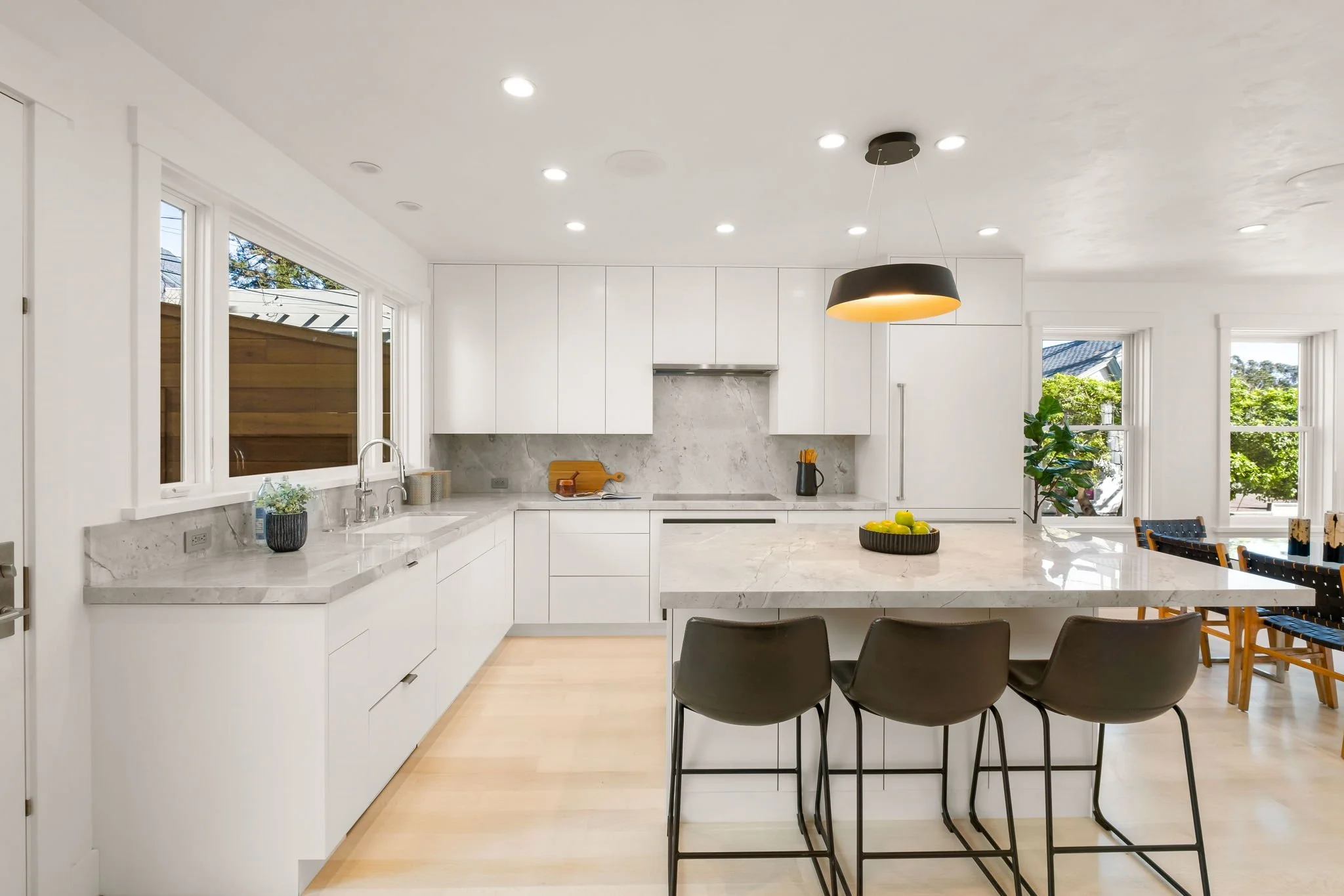 Modern white kitchen with marble countertops, black pendant light, three black barstools at a kitchen island, and large windows with views of trees and a neighboring house.