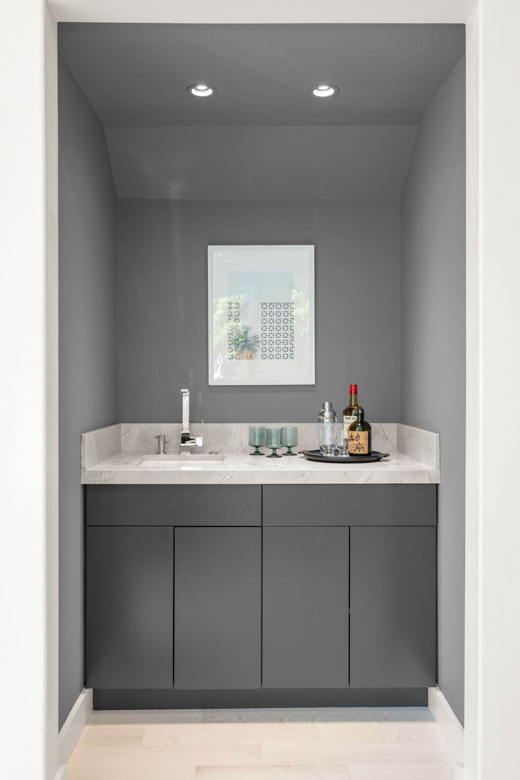 Small modern bar nook with gray cabinetry, marble countertop, and a framed mirror. Items on the counter include three green glasses, a cocktail shaker, a liquor bottle, and a tray with a drink.