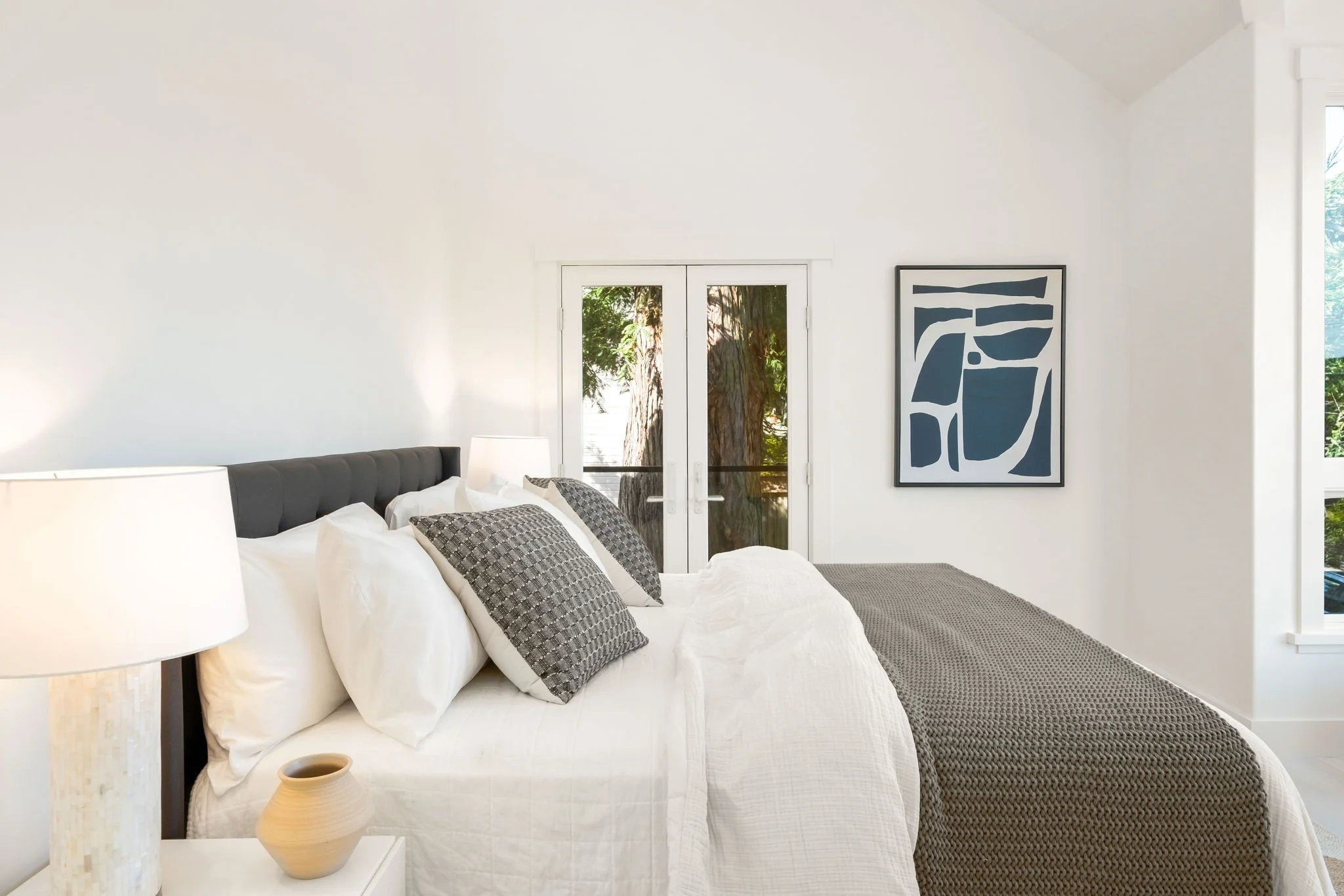 A bright bedroom with a large bed, white bedding, gray pillows, and a dark gray headboard. There are two lamps on white nightstands and a small beige vase on the left. The room has a glass door with a view of greenery outside and a framed abstract ar