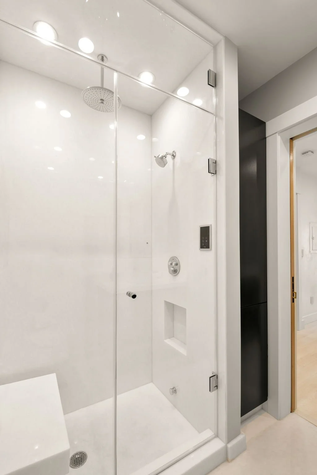 Modern walk-in shower with white walls and ceiling, glass door, overhead rainfall showerhead, wall-mounted showerhead, digital control panel, small recessed shelf, and black storage cabinet nearby.