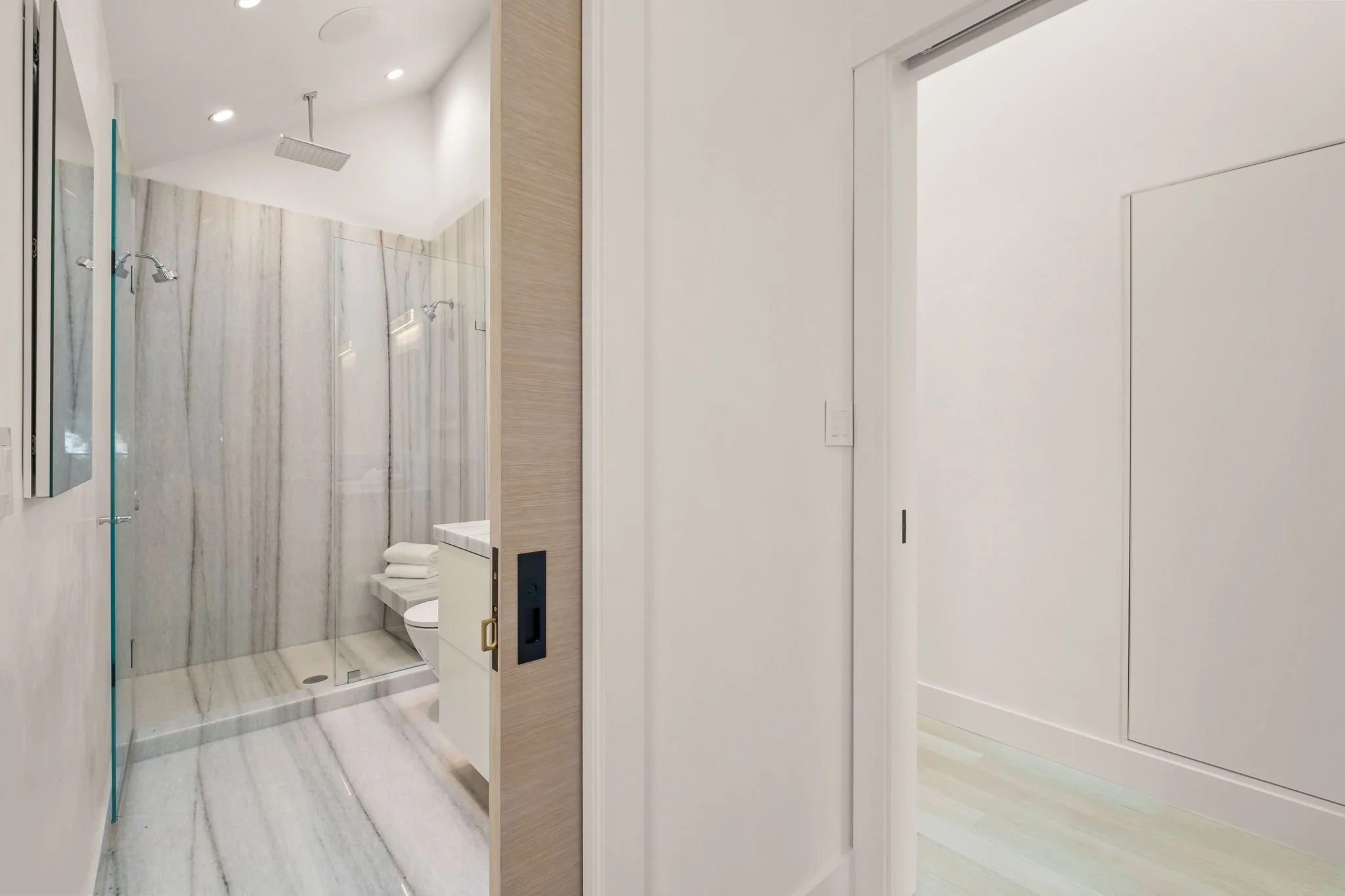 Modern bathroom with marble shower walls, rain showerheads, a toilet, and a small vanity with towels, viewed through an open door in a minimalist white room.