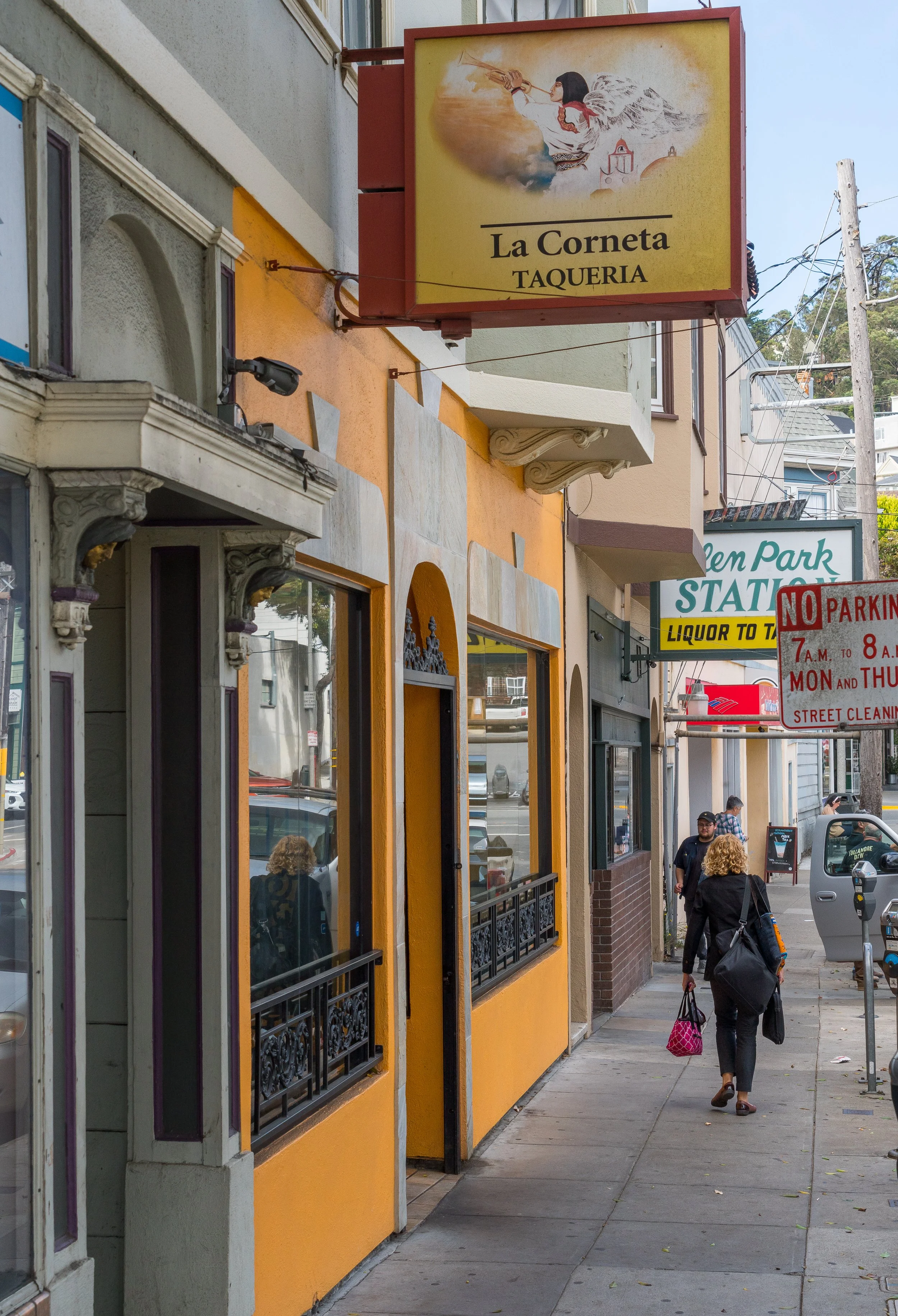 Street view with colorful buildings, signs, and pedestrians. Visible signs include 'La Corneta Taqueria' and a parking restriction notice. People walk on the sidewalk next to parked vehicles.