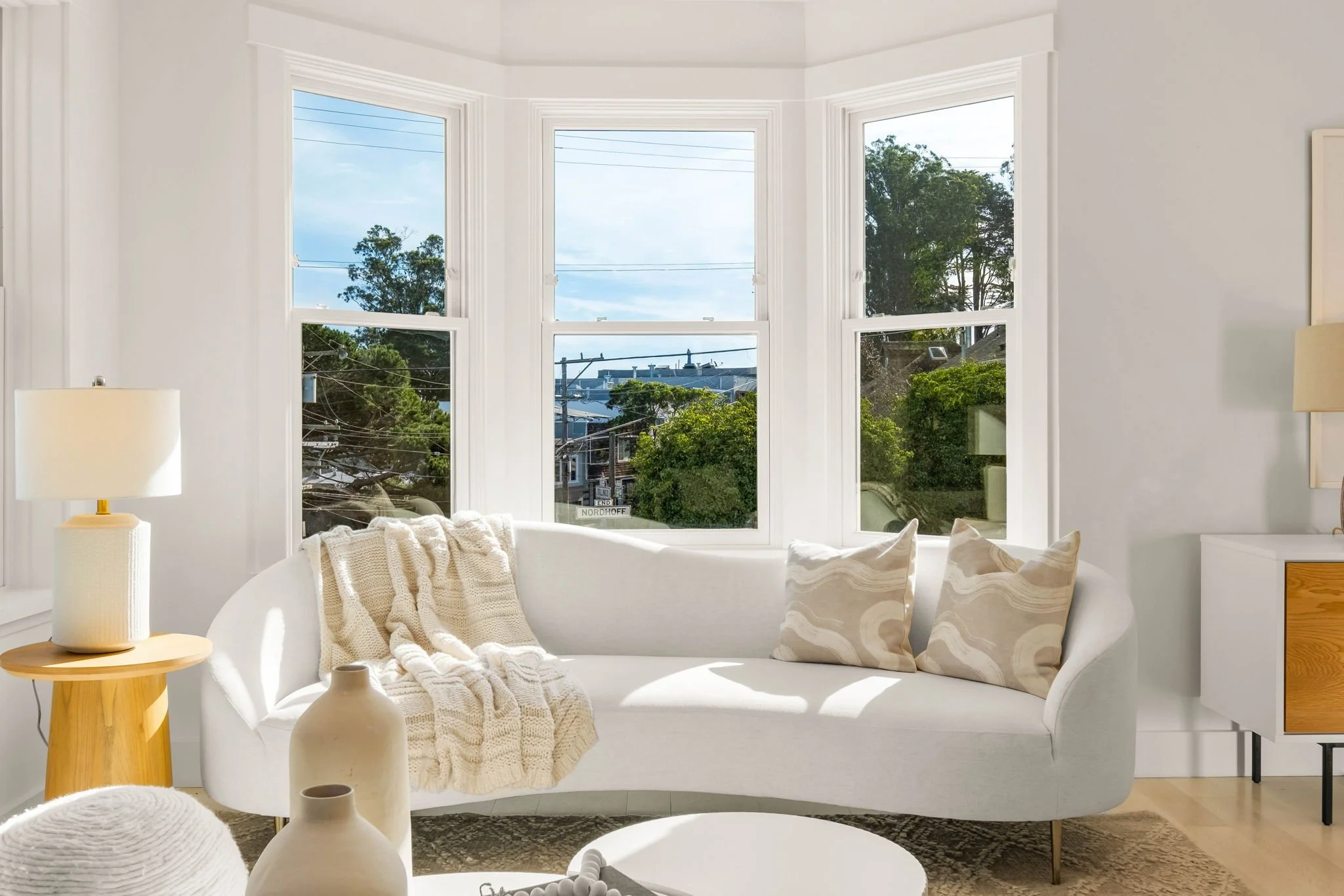 Bright living room with a white curved sofa, beige and white pillows, cream throw blanket, round white coffee table, wooden side table with lamp, large bay windows showing trees and a neighborhood view.