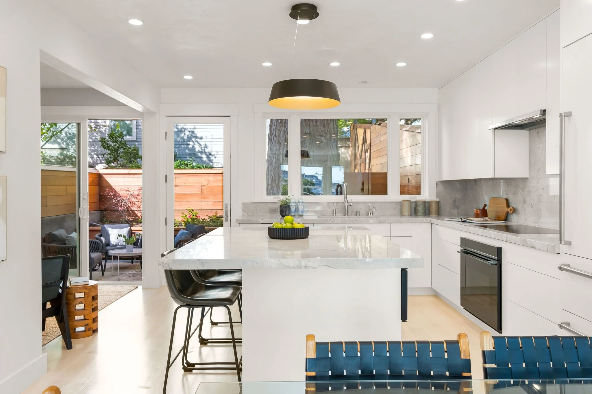Modern white kitchen with marble countertops, black pendant light, and sliding glass door leading to outdoor patio.
