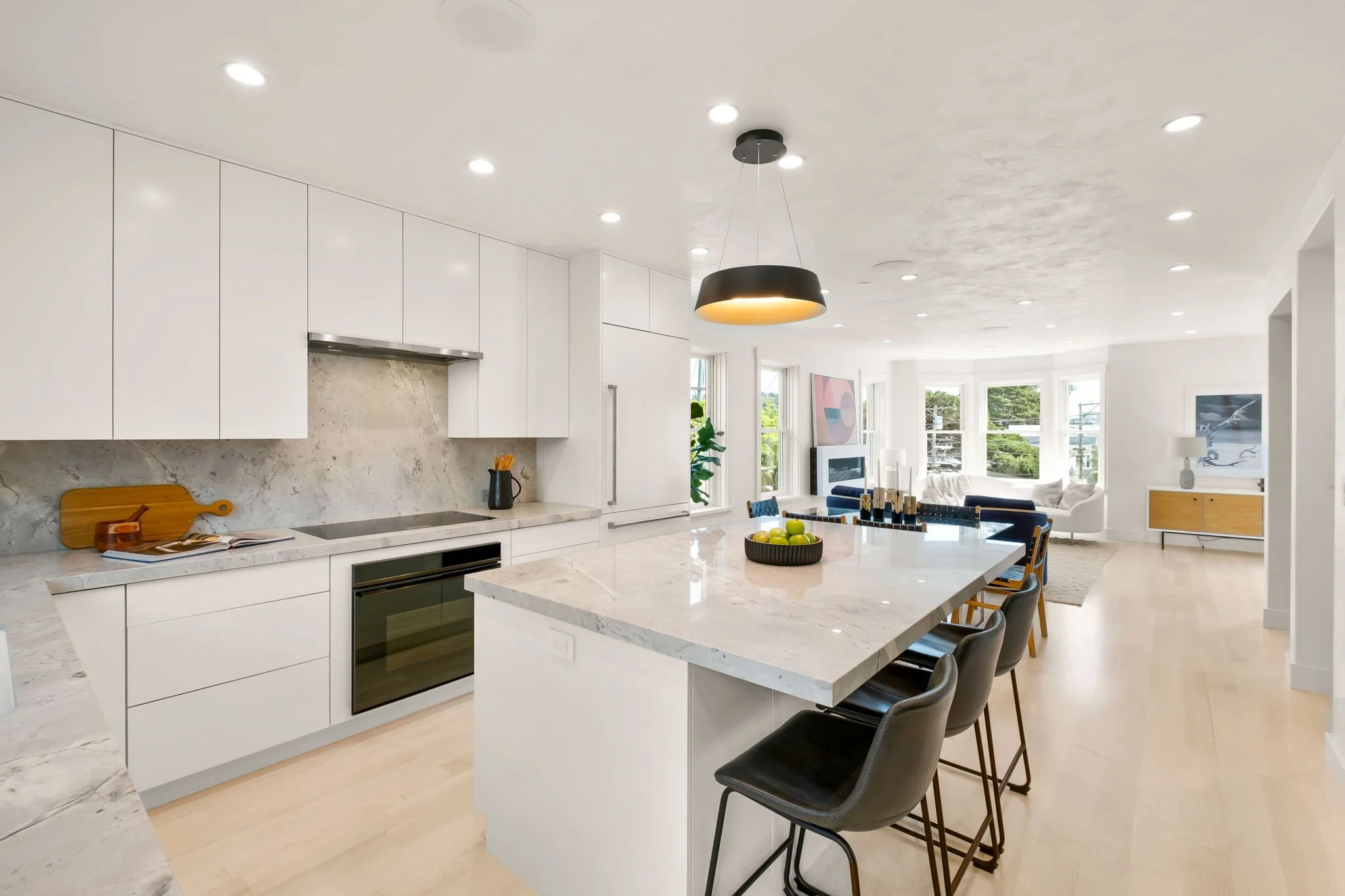 Modern open-concept kitchen and living room with white cabinetry, marble countertops, a central island with seating, black pendant light, and large windows in the background.