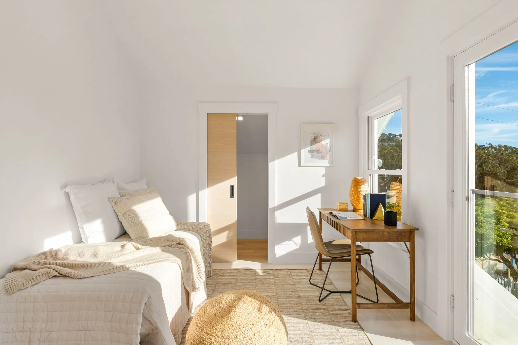 Bright and airy bedroom with a white bed, beige pillow, knitted throw blanket, wooden desk with decorative items, large windows showing trees outside, and sunlight streaming into the room.