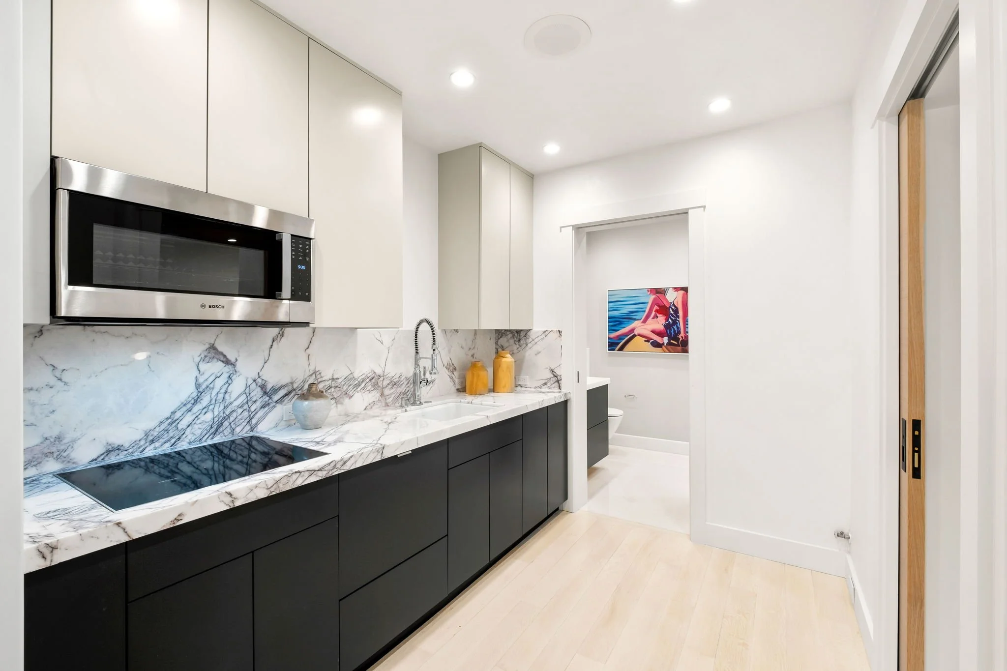 Modern kitchen with white upper cabinets, black lower cabinets, marble countertop and backsplash, stainless steel microwave, and a sink. There is a doorway at the end leading to a room with a framed painting of a boat on water.