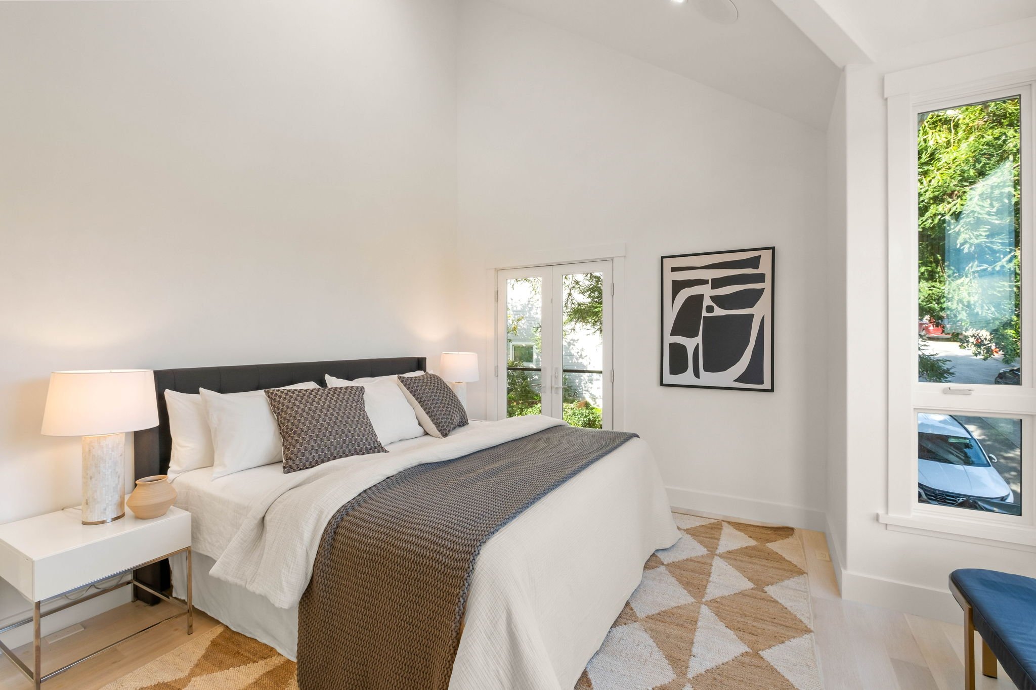 Bright bedroom with white walls, large bed with white bedding, gray headboard, and two pillows with patterned pillowcases. Two bedside lamps on white side tables, artwork on wall, large window showing greenery, and a patterned rug on light wood floor