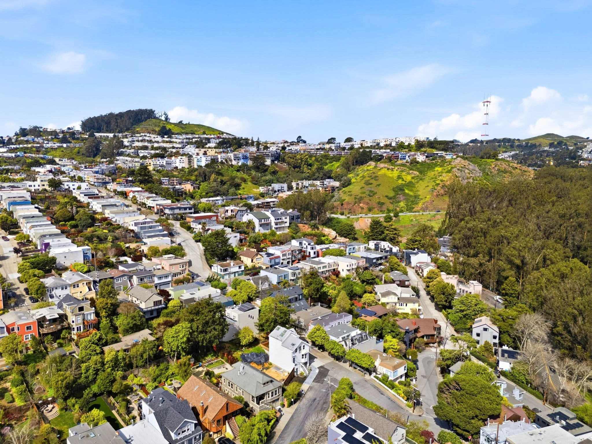 Aerial view of a hilly residential neighborhood with colorful houses, trees, and greenery under a partly cloudy sky.