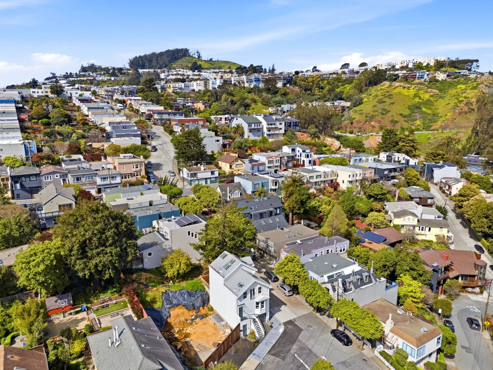 Aerial view of a hillside residential neighborhood with a mix of houses, trees, and winding streets, with a green hill in the background and a partly cloudy sky.