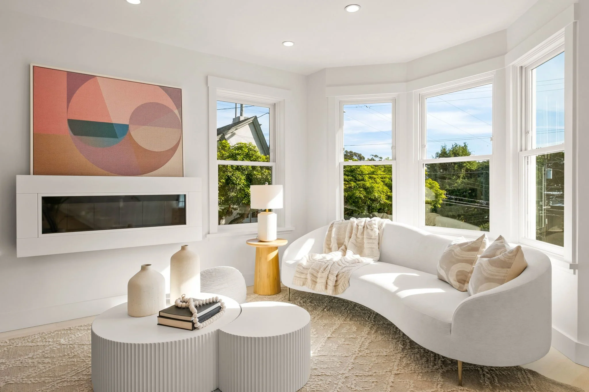 Bright living room with large windows, white curved sofa with pillows, round white coffee tables, decorative vases, and abstract wall art