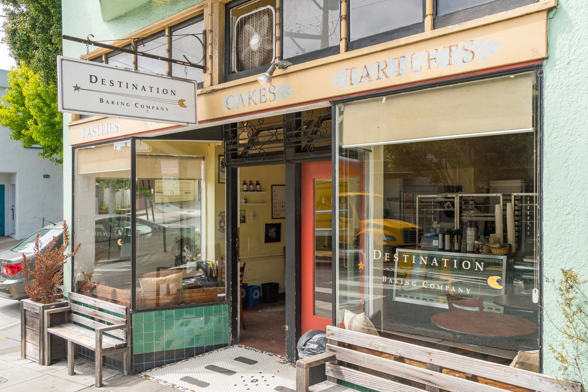 Exterior view of a bakery called 'Destination Baking Company' with glass windows and colorful signage, including words like 'Pastries,' 'Cakes,' and 'Tartlets', benches outside, and cars parked on the street.