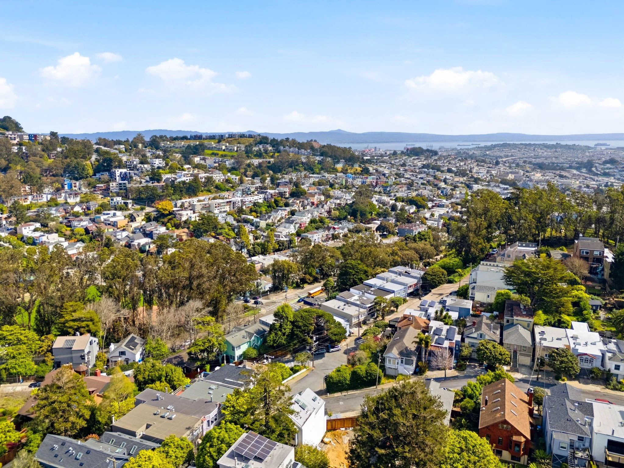 Aerial view of a densely populated coastal city with residential houses, greenery, and hills under a blue sky with clouds.