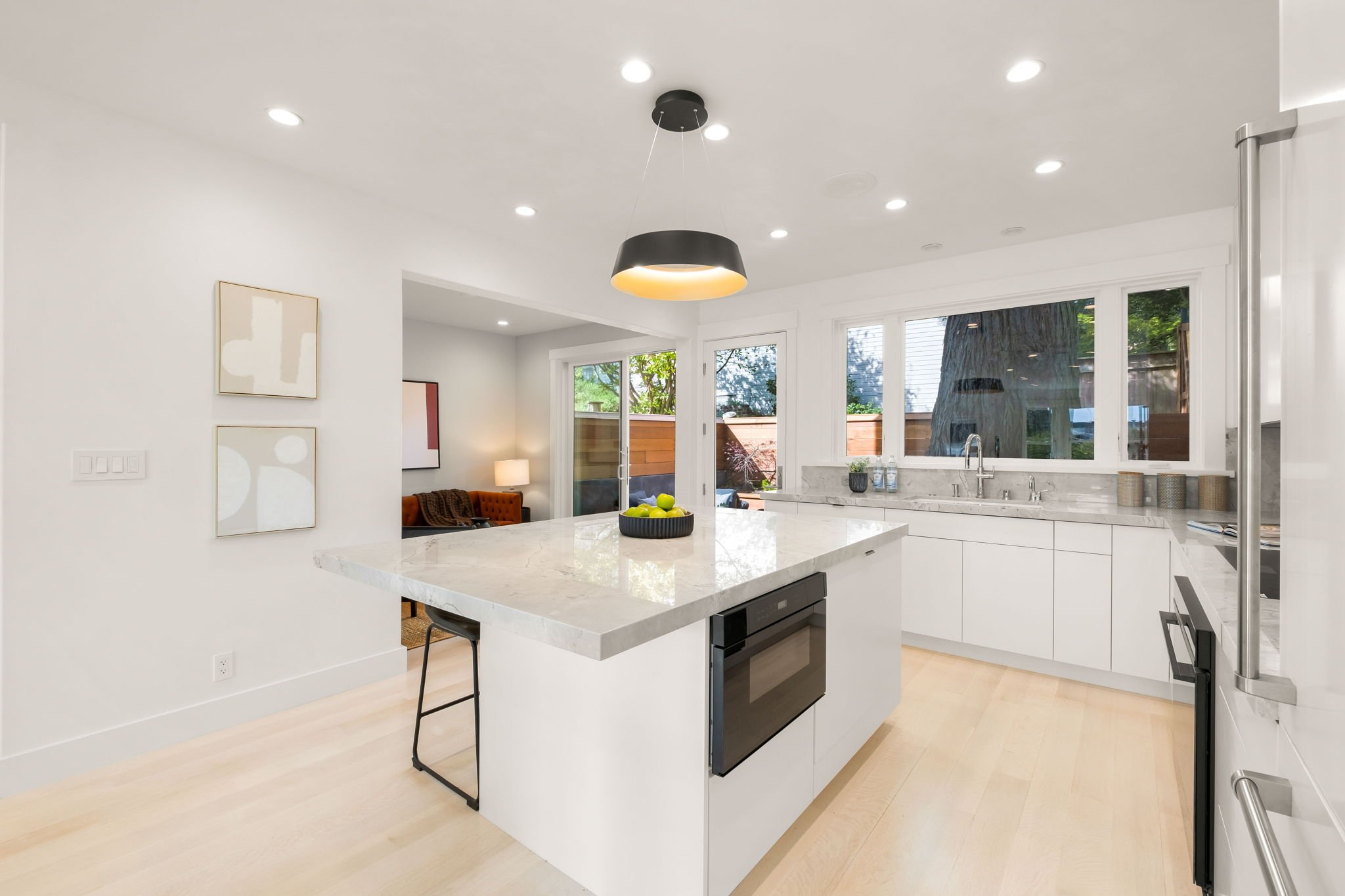 Modern kitchen with white cabinets, marble countertops, large island with a black bowl of green apples, built-in oven, stainless steel refrigerator, large windows, and a view into a living room with a brown sofa and paintings.