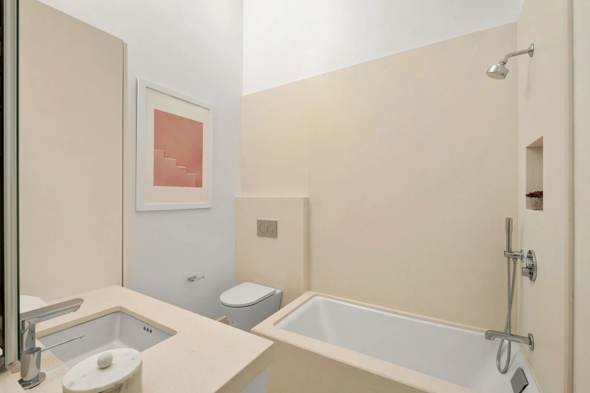 A clean, modern bathroom with a vanity sink, framed pink artwork, a toilet, a bathtub with a shower, and neutral-colored walls.