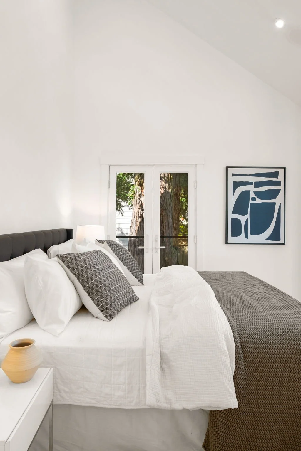 A neatly made bed with white and patterned pillows in a bright bedroom. A window with views of trees, and a framed abstract art piece on the wall.