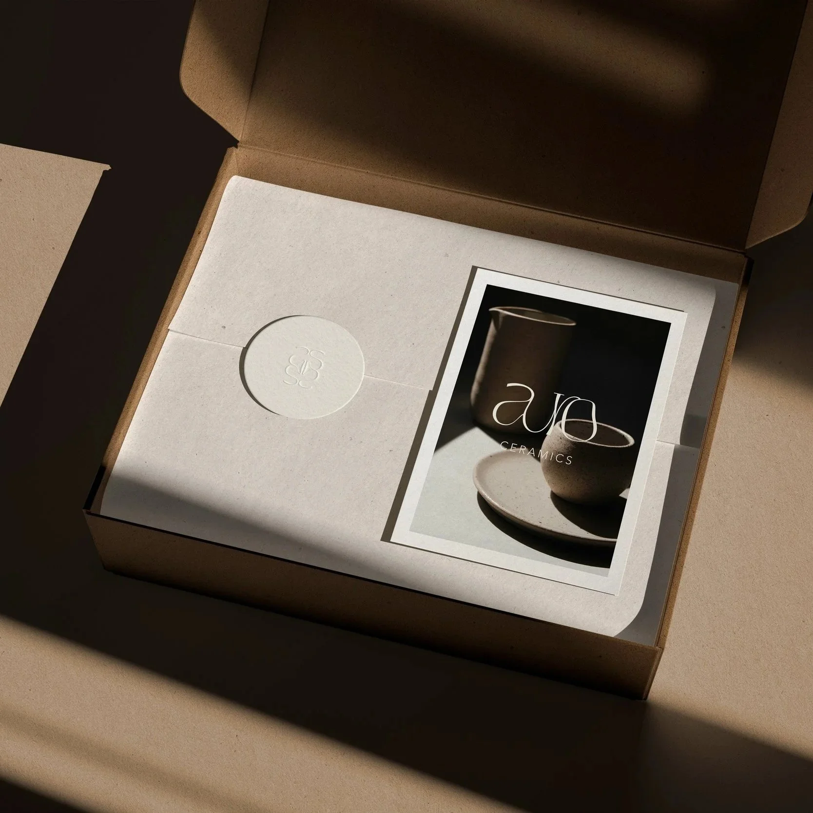 Open cardboard box containing a white envelope with an embossed logo and a brochure showing ceramics, including a pitcher and bowls, with a simple logo 'Auro CERAMICS' on a photograph.
