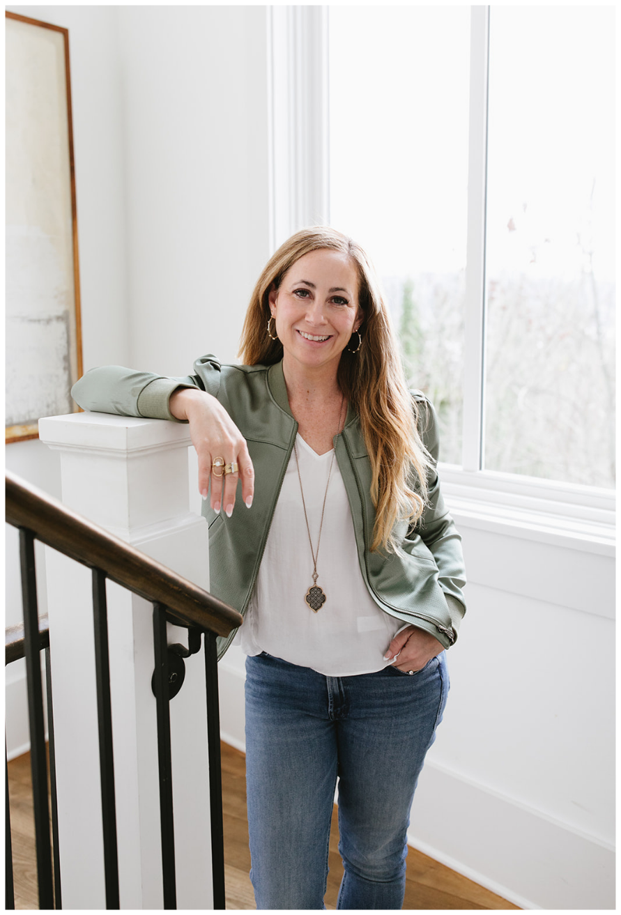 Tara Townsend Seattle real estate agent standing by staircase in light-filled home, wearing green jacket and casual style