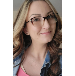 Close-up of a young woman with long wavy brown hair wearing glasses, a pink top, and a denim jacket, smiling at the camera.
