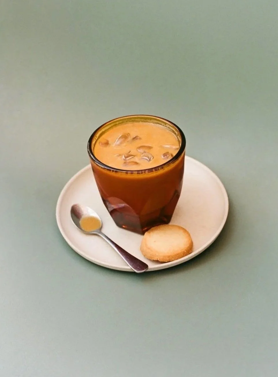 A glass of iced tea on a white plate with a slice of bread, a teaspoon, and a bagel chip on a light green surface