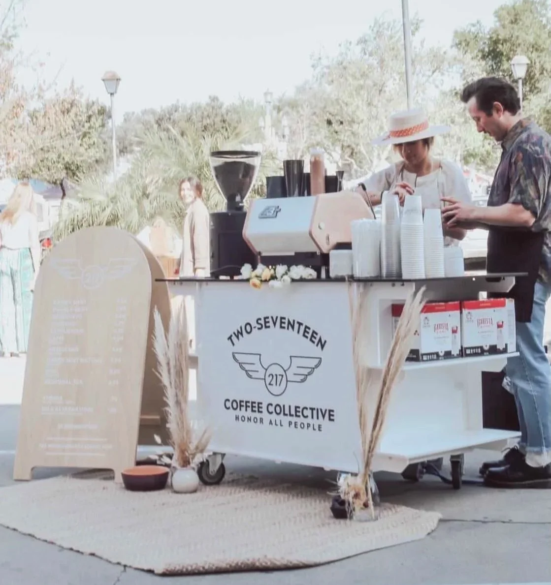 A small outdoor coffee cart with two people serving coffee, surrounded by trees and festival-goers, with a sign reading "Two-Seventeen Coffee Collective" and a menu board.