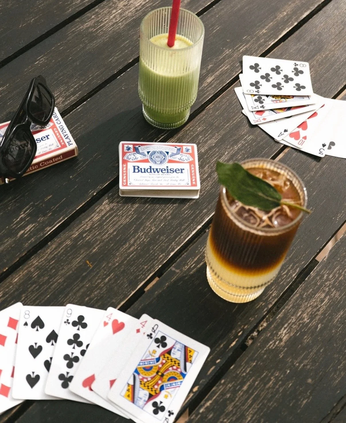 A table set with playing cards, two drinks, a pack of matches, a pack of cigarettes, a pair of sunglasses, and a deck of beer coasters.