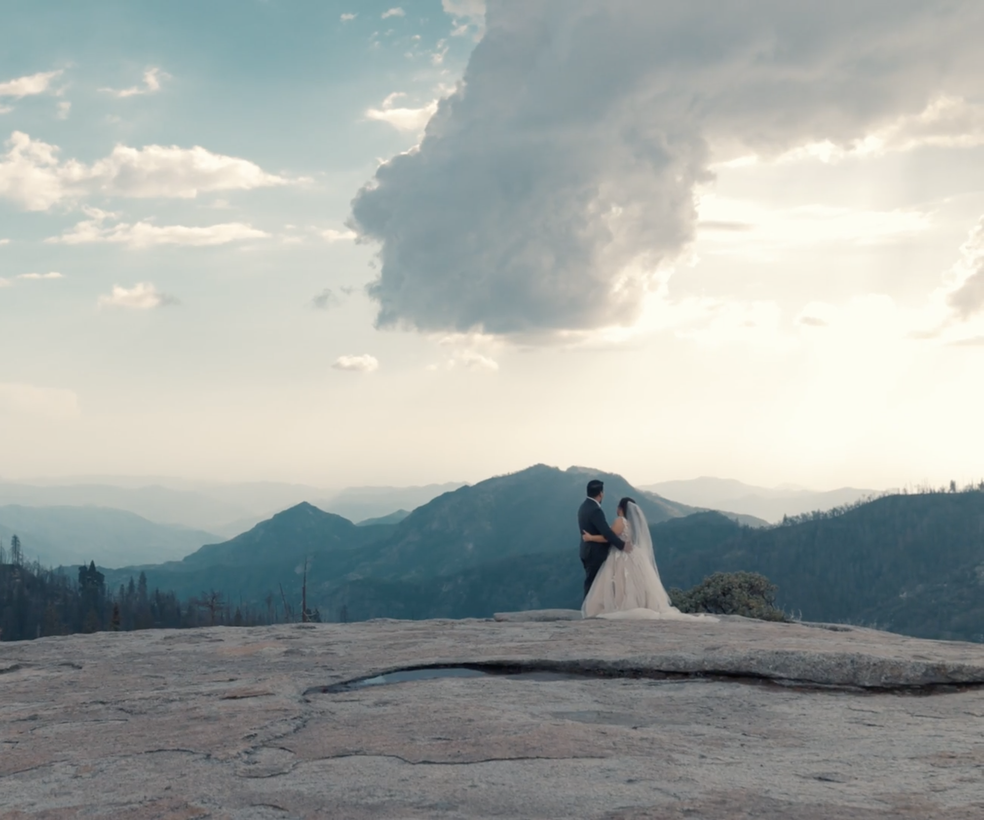 A bride and groom standing close on top of a rocky hill with panoramic mountain views in the background, under a partly cloudy sky with the sun shining.