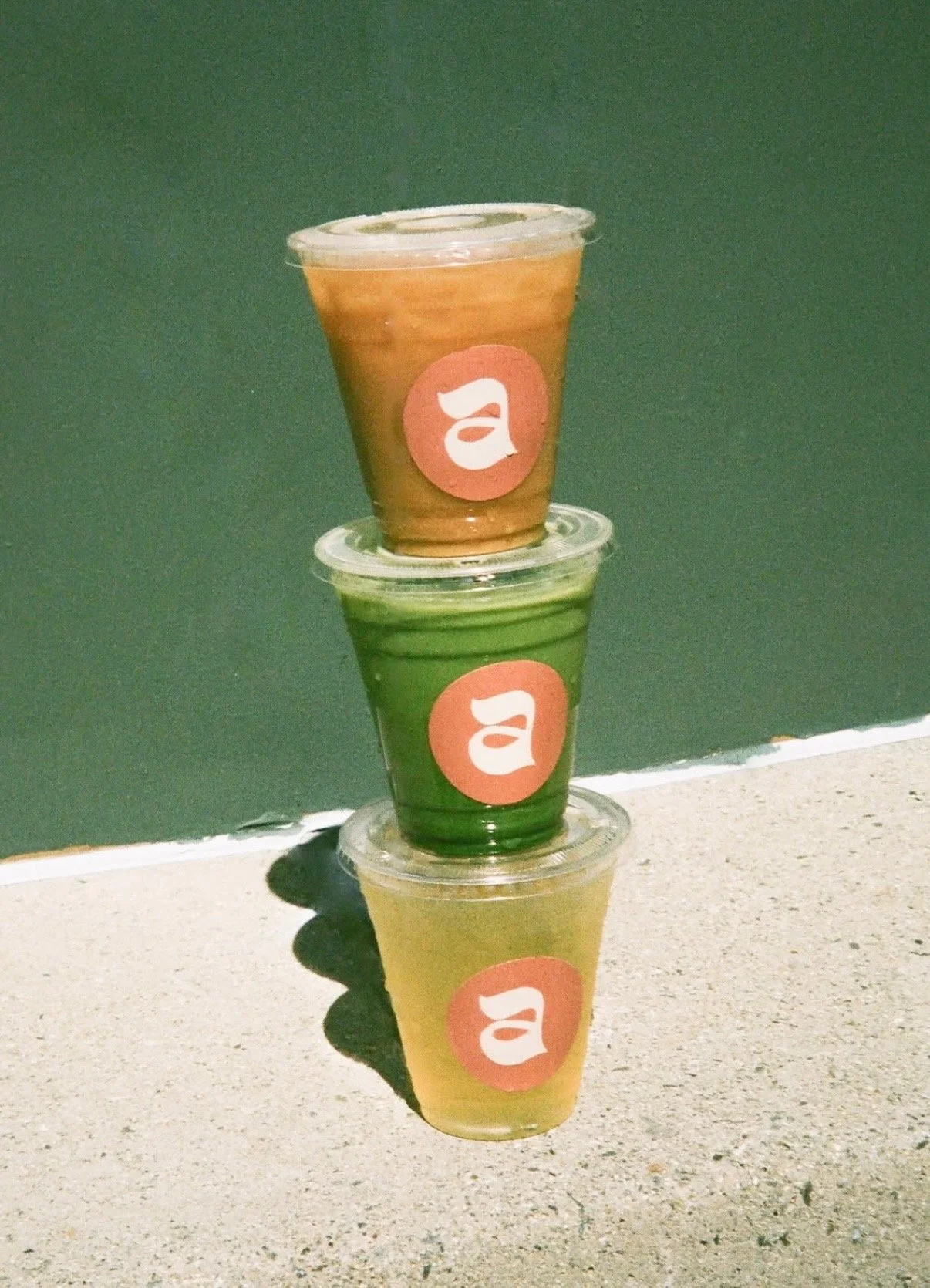 Three clear plastic cups filled with different colored beverages stacked on top of each other, with a red circle and white letter 'a' logo on each cup, placed on a light-colored surface next to a green wall.