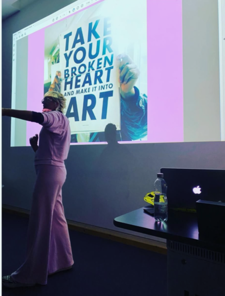 A woman standing in front of a large presentation screen displaying the text 'Take your broken heart and make it into art'. She is pointing towards the screen, wearing purple clothing, in a classroom or conference room setting with a laptop and water bottle on a table nearby.