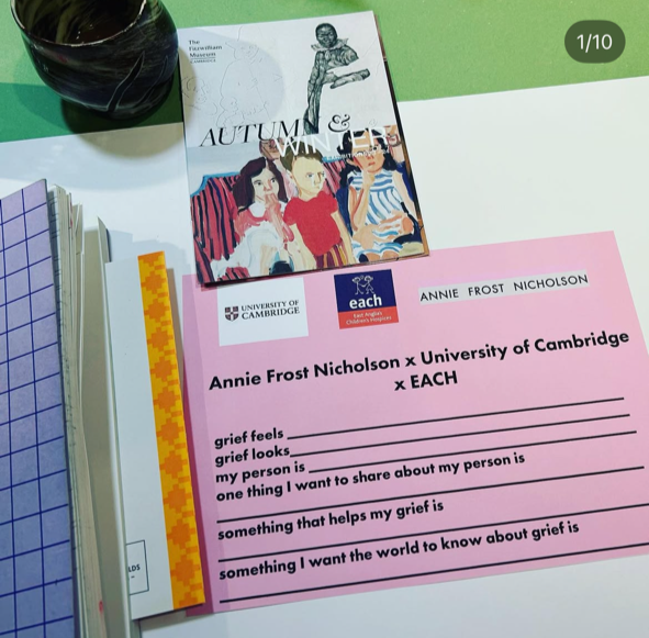 A pink worksheet titled 'Annie Frost Nicholson x University of Cambridge x EACH,' with prompts about grief feelings and sharing, placed on a desk with a purple notebook, a strip of yellow washi tape, a book titled 'Autumn,' a black coffee mug, and some papers.