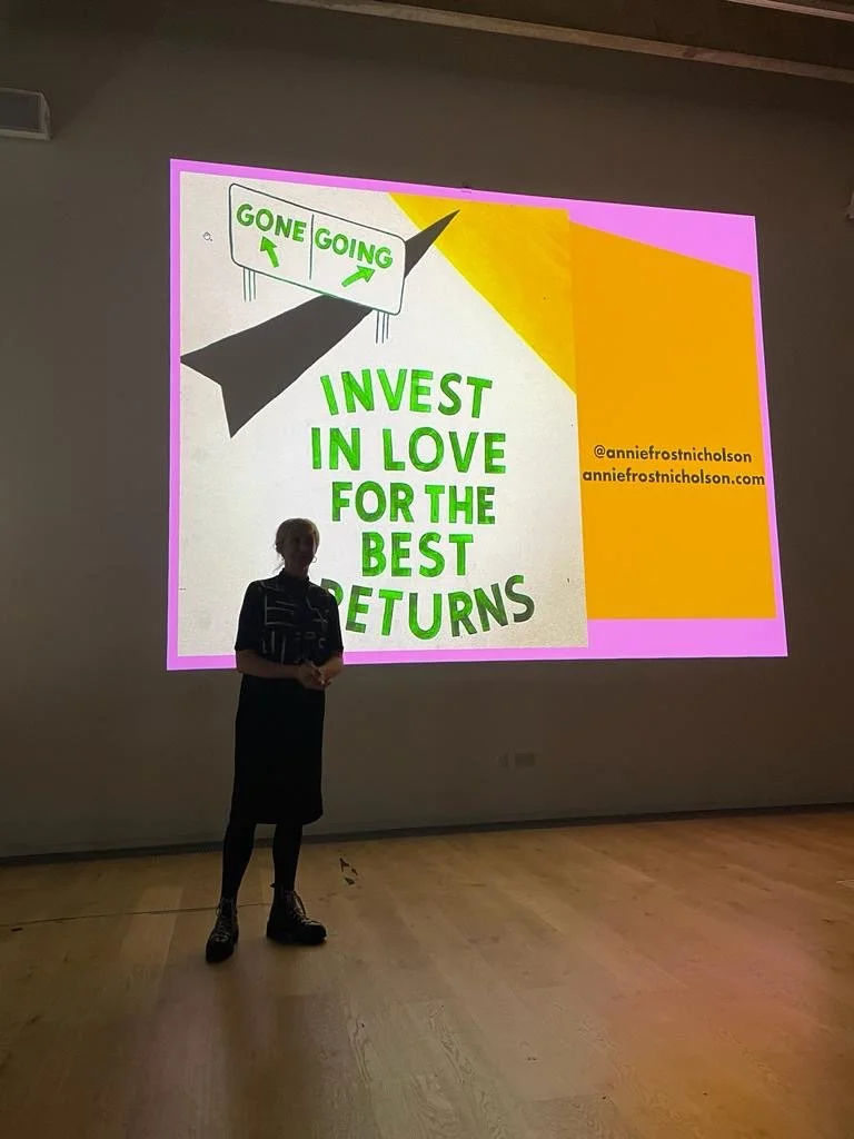A person standing in front of a presentation slide that says, "Invest in love for the best returns." The slide includes a graphic of a sign with the words "GONE" and "GOING" and an arrow, along with social media handles and a website link.