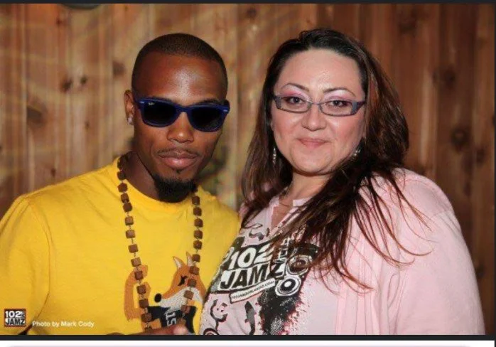 Two people posing for a photo indoors, with a wooden wall in the background. The person on the left is a man wearing sunglasses, a yellow shirt, and a beaded necklace. The person on the right is a woman with long dark hair, wearing glasses and a pink
