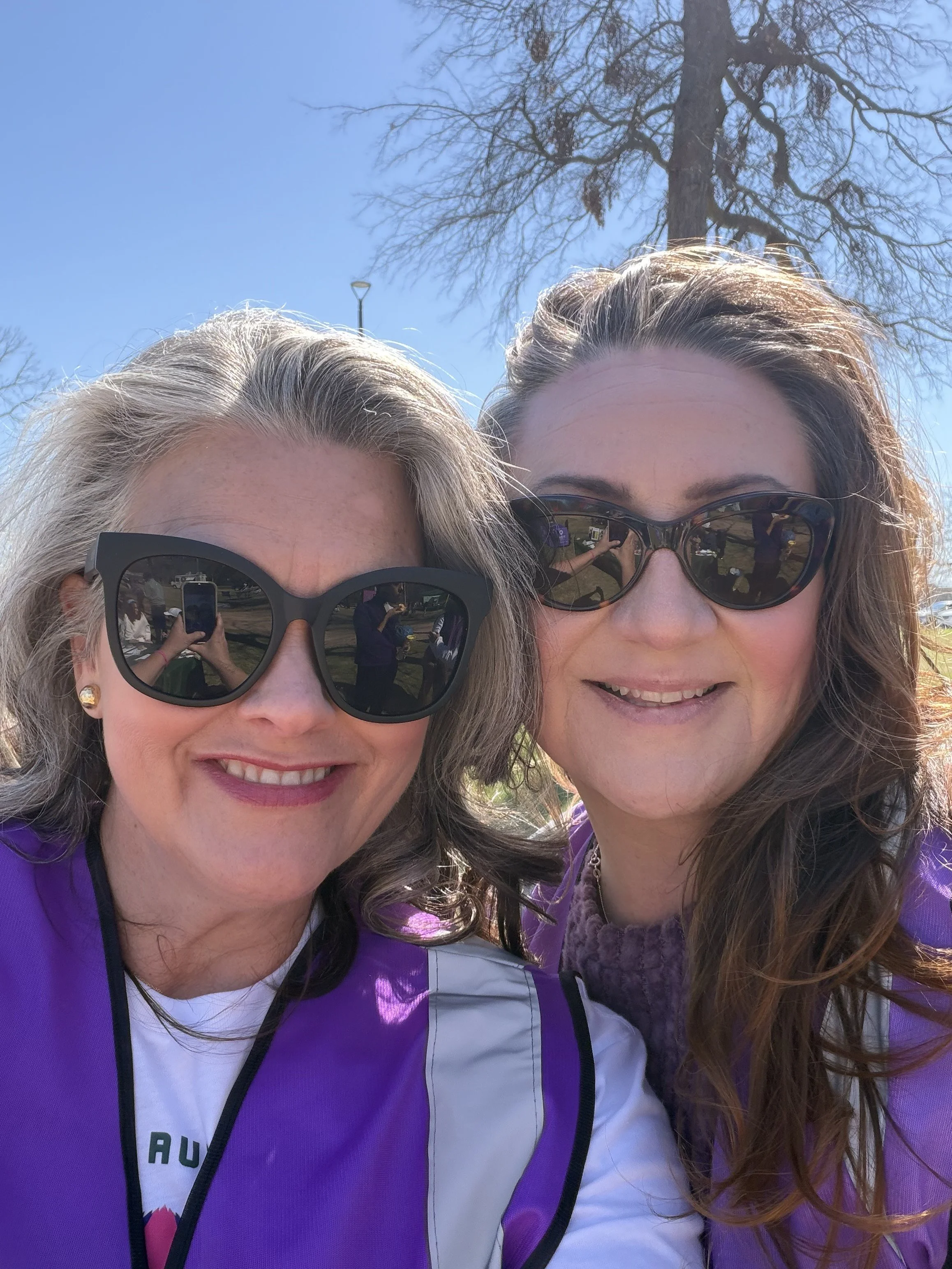 Two women wearing sunglasses and purple jackets smiling for a selfie outdoors on a sunny day, with a tree and blue sky in the background.