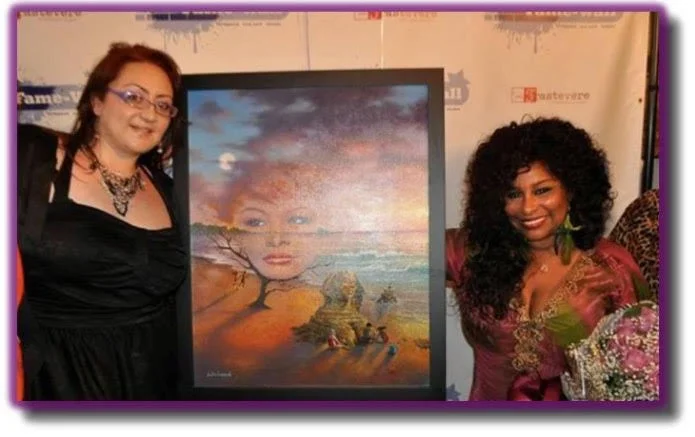 Two women standing beside a framed painting at an event. The painting features a woman's face, a tree, and a landscape scene with a boat.