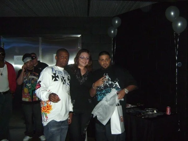 Group of people at a celebration, some holding drinks, with balloons and a DJ setup in a dimly lit room.