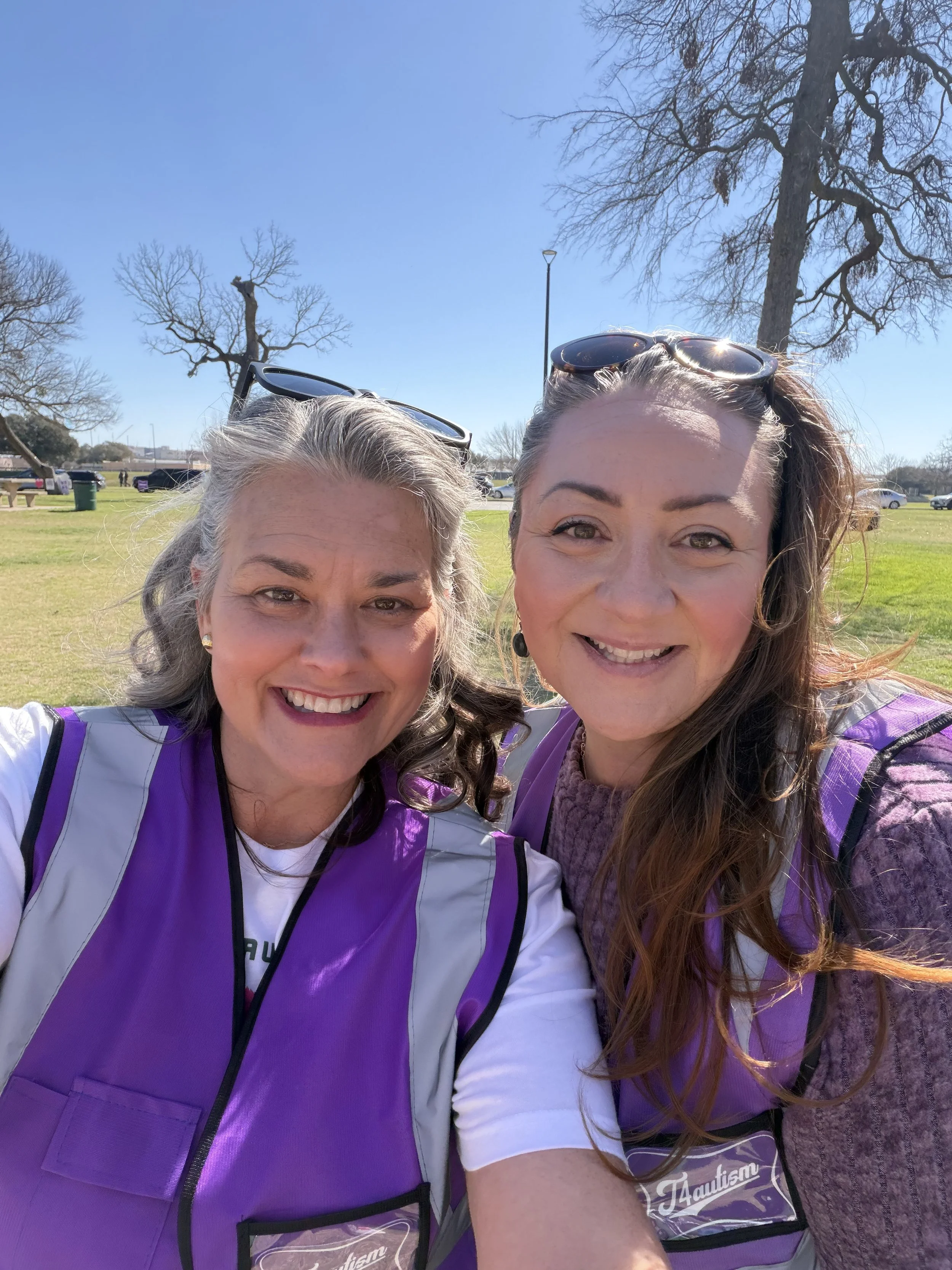 Two women smiling outdoors on a sunny day, wearing purple vests with a logo, with leafless trees and a grassy field in the background.