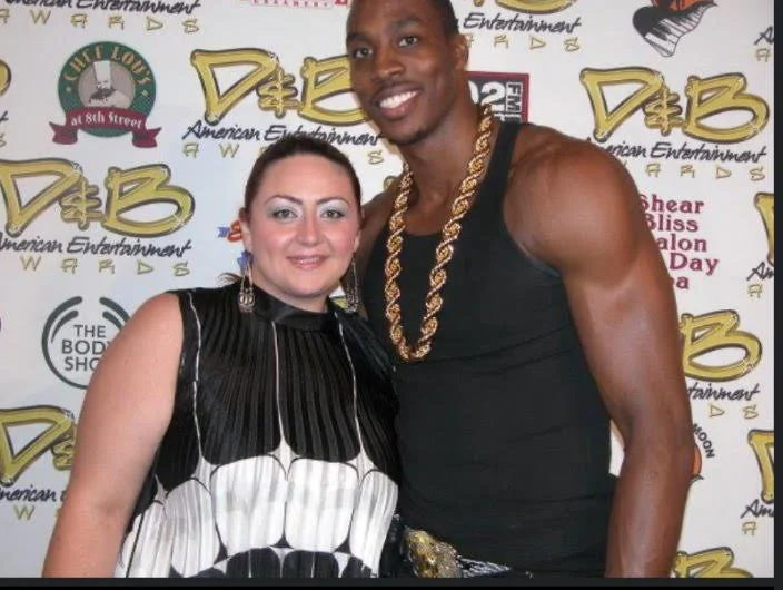 A smiling woman and a muscular man wearing a gold chain pose together at an event with a backdrop featuring the D&B logo and various text and logos related to American entertainment.