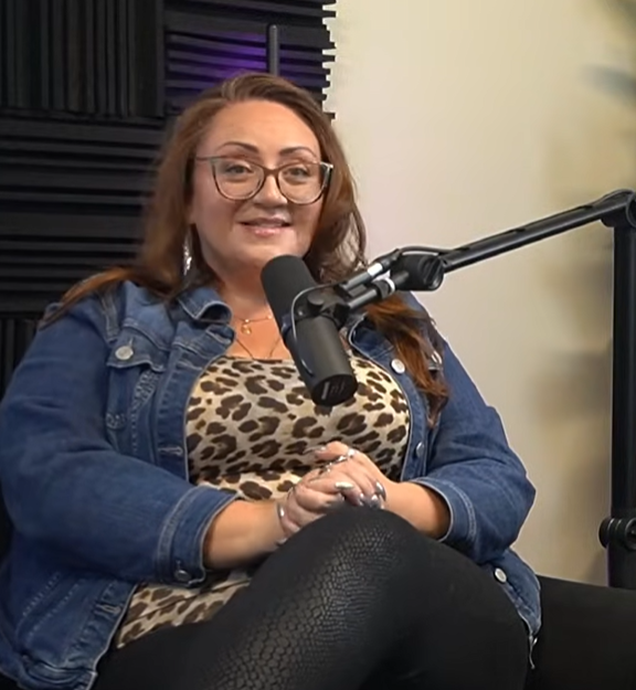 A woman with glasses and long red hair, wearing a leopard print top and denim jacket, sitting in front of a microphone in a recording studio.