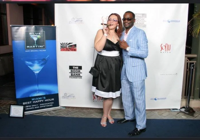 A woman and man in formal attire posing together at a promotional event, with a backdrop displaying logos and a blue martini sign.