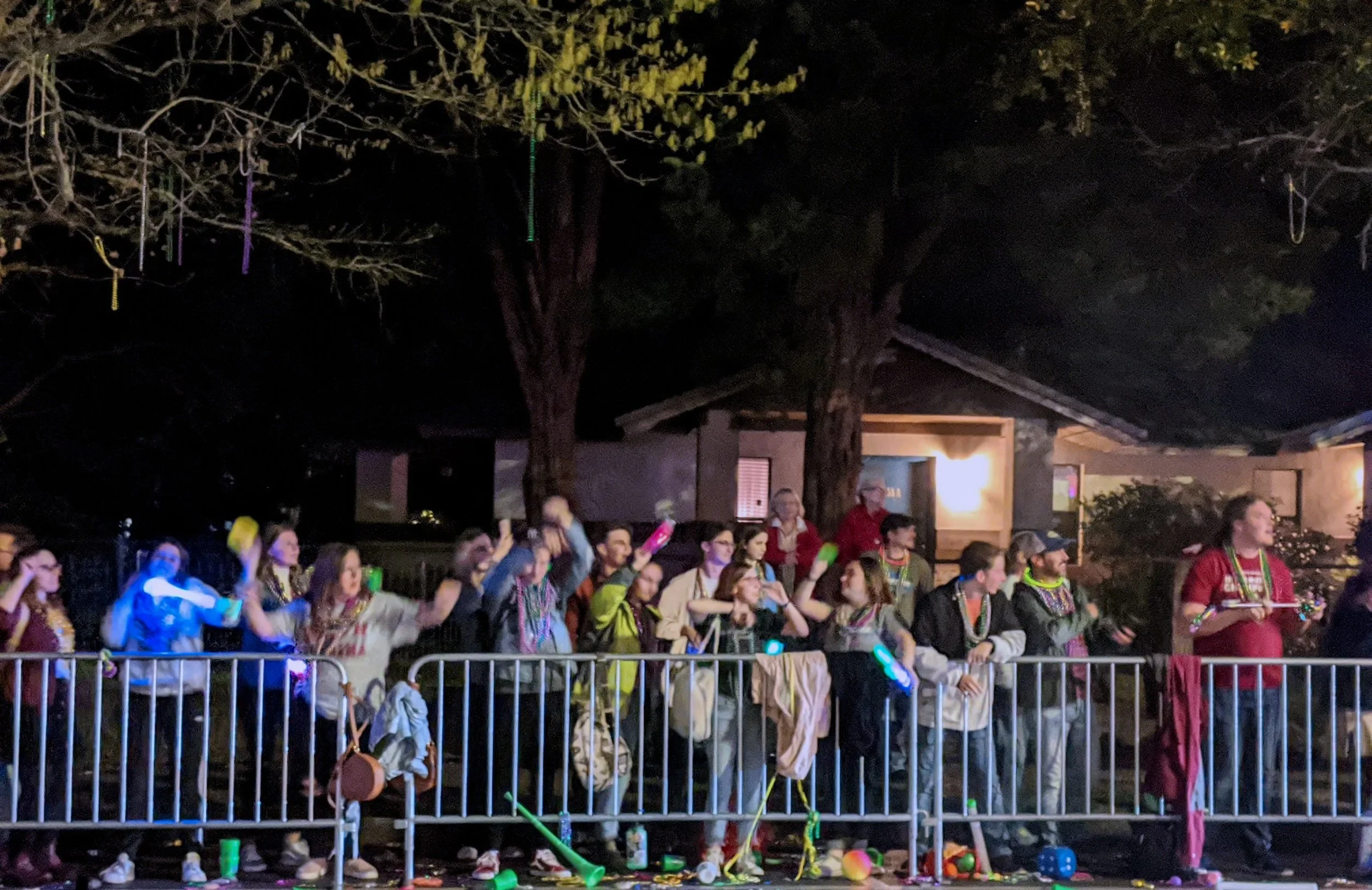 Group of people standing behind a barrier at night, some wearing glow necklaces and holding glow sticks, with trees and a house in the background.