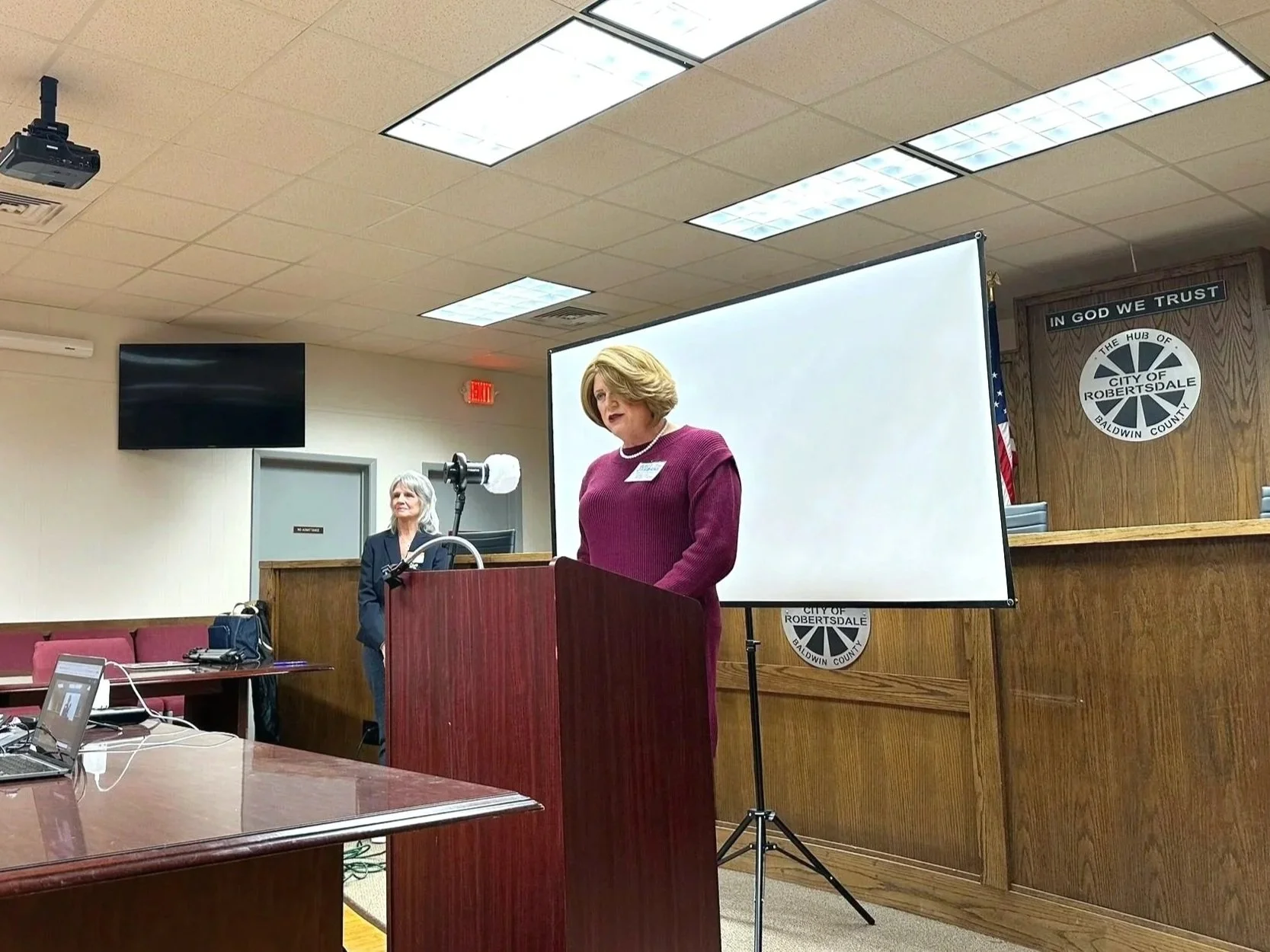 A woman in a purple dress is standing at a wooden podium in a room, speaking into a microphone. Behind her, there is a large white screen, a woman with gray hair and glasses standing near a camera, and an official seal on the wall that reads "City of Robertsdale Baldwin County" along with an American flag.
