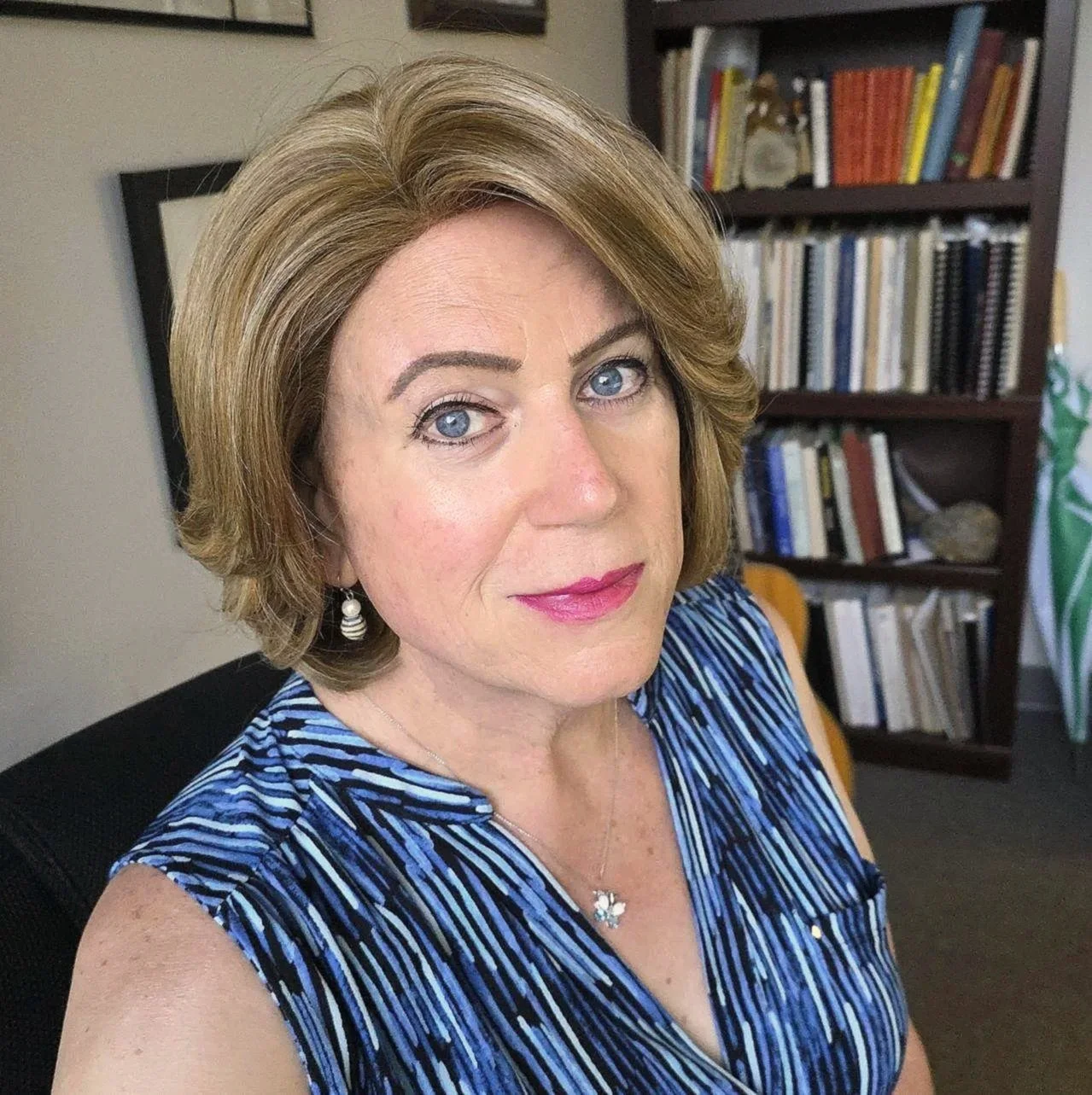 A woman with shoulder-length light brown hair and blue eyes, wearing a striped blue and black sleeveless top, earrings, and a butterfly necklace, sitting in an office space with a bookshelf filled with books in the background.