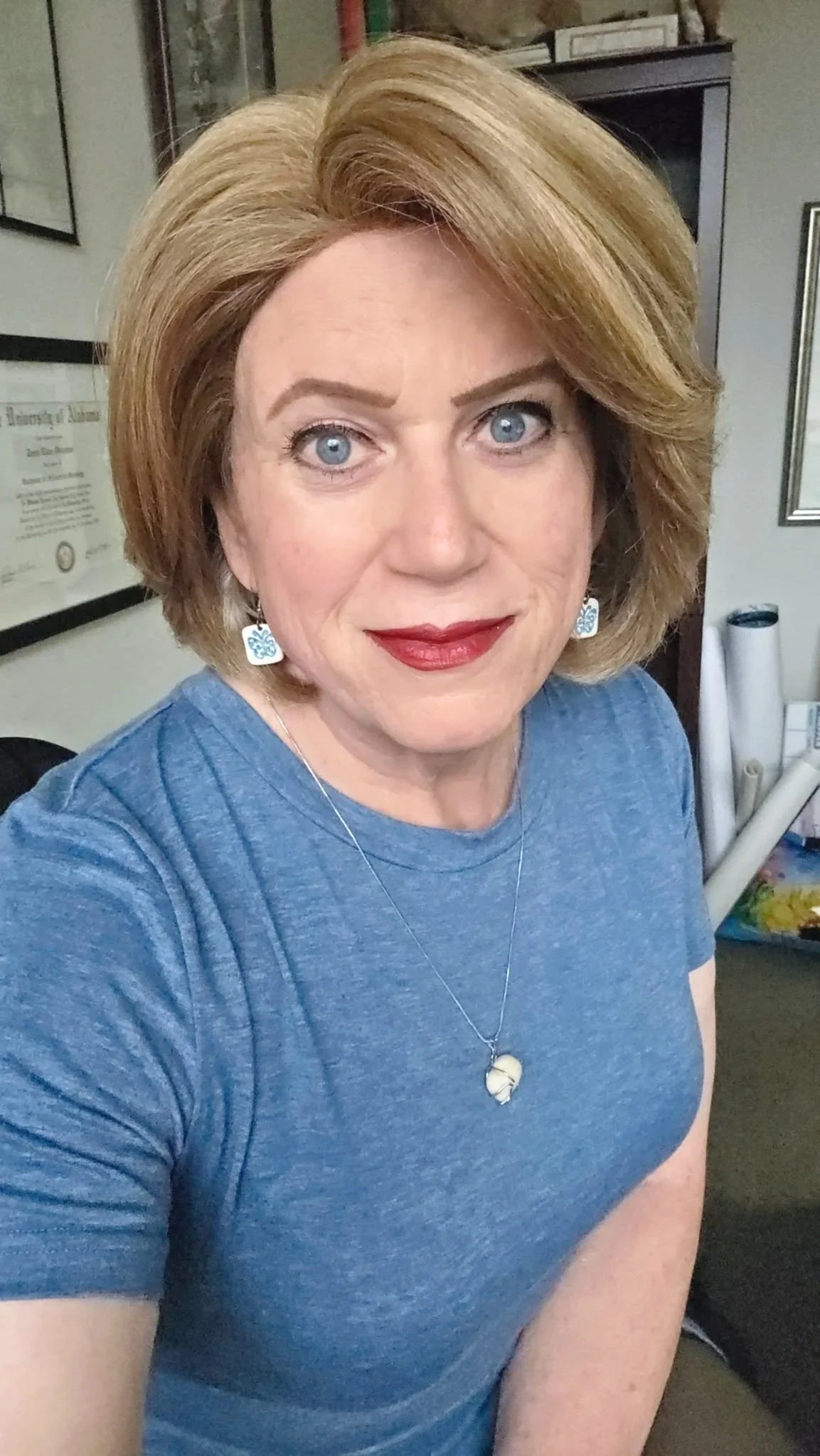 A woman with short blonde hair, blue eyes, wearing a blue t-shirt, earrings, and a necklace, standing in an office or workspace with framed certificates and rolled-up posters in the background.